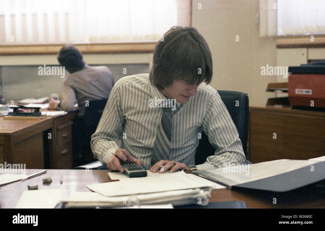 1970s, historical, a male, late twenties, or early thirties in age in an office, sitting at a desk, working, a financial manager or possibly an accountant as he is using a small calculator to add figures from printed ledgers or journals, England, UK. Stock Photo