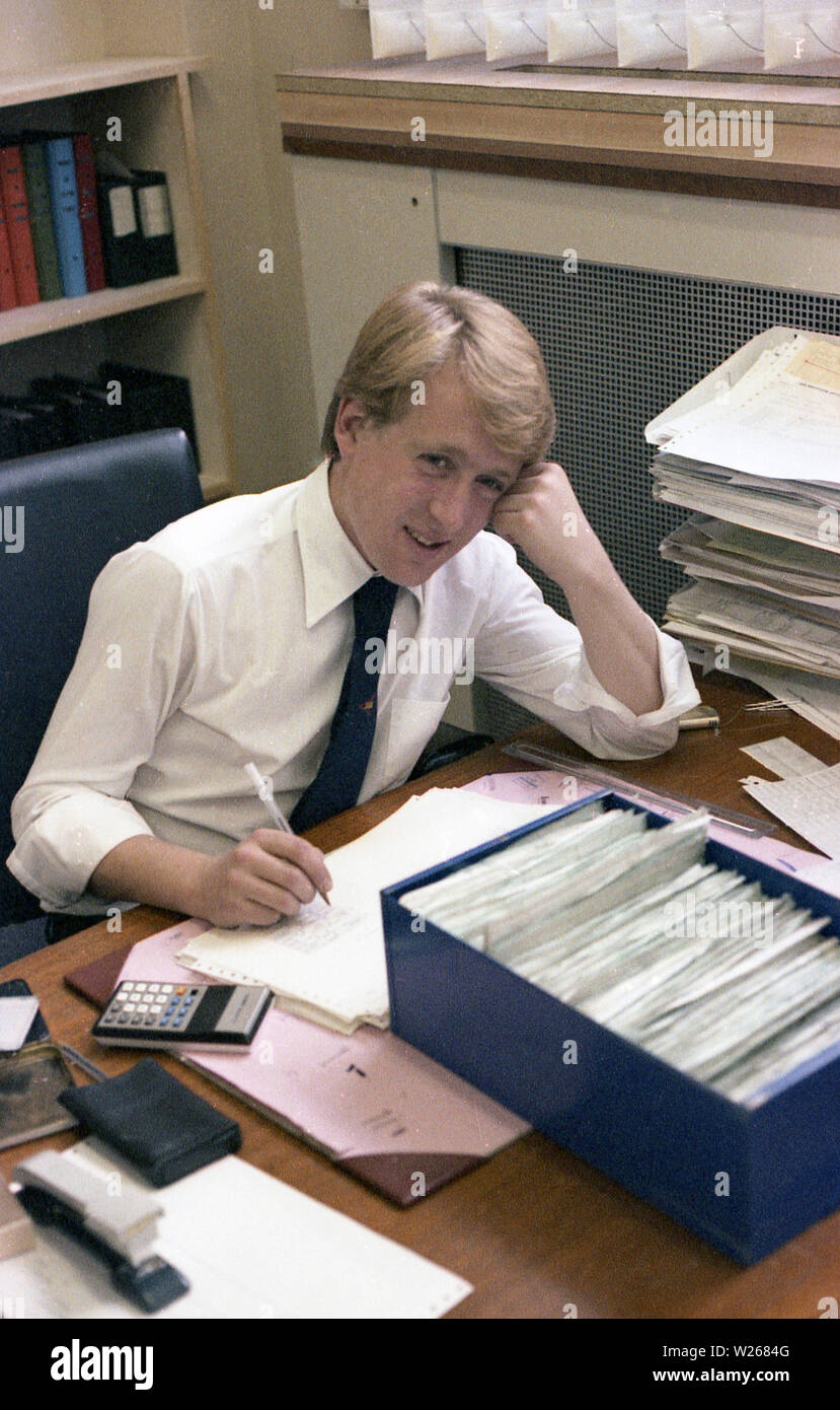 1970s, historical, a young adult male, late twenties, or early thirties in age in an office, sitting at a desk, working, a financial manager or possibly an accountant as he is using a calculator to add figures from printed ledgers or journals, England, UK. Stock Photo