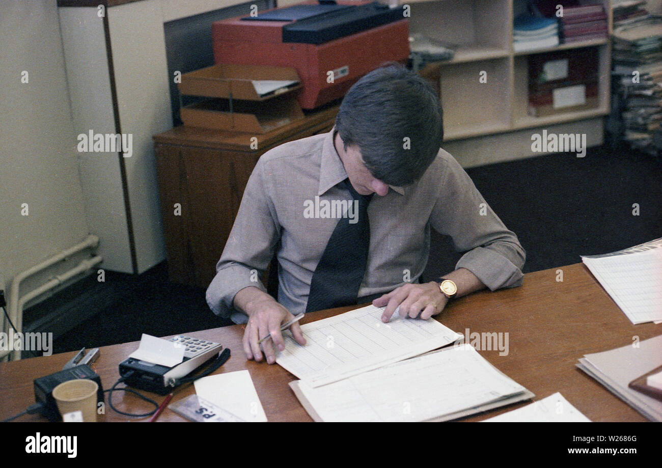 1970s, historical, a male, late twenties, or early thirties in age in an office, sitting at a desk, working, a financial manager or possibly an accountant as he is using a small electronic calculator to add figures from printed ledgers or journals, England, UK. Stock Photo