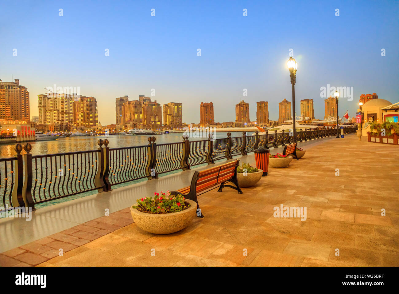 Benches along marina corniche walkway in Porto Arabia at the Pearl-Qatar, Doha, with residential skyscrapers of artificial island illuminated at night Stock Photo