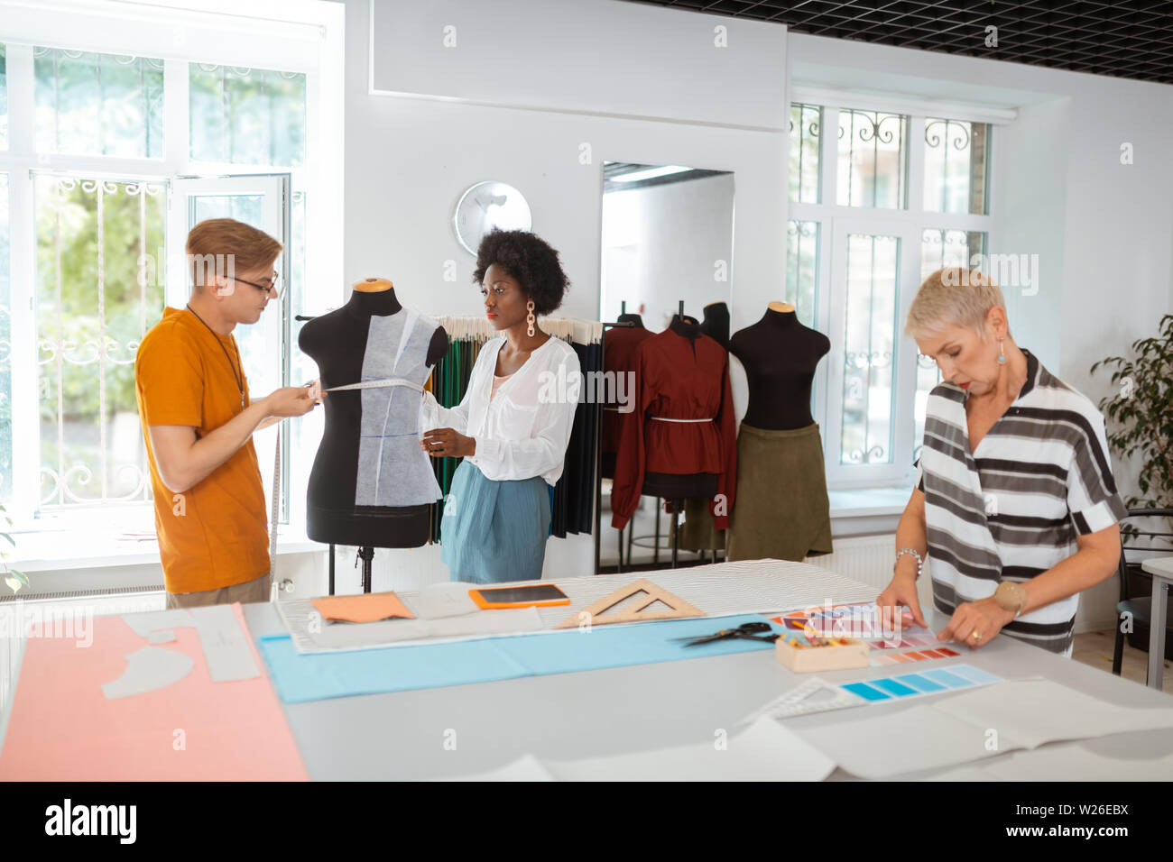 Creative team. Two young fashion designers taking measurements on the tailor dummy while their senior coworker choosing the color palette Stock Photo