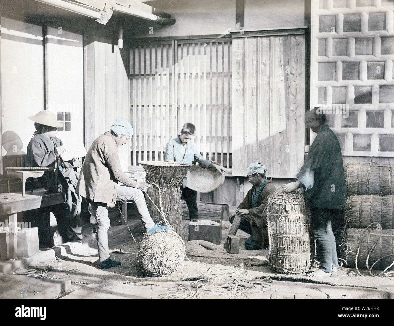 [ 1880s Japan - Packing Rice ] —   Two men in the foreground are packing rice in straw bags, while the man on the left is taking records. A soroban (Japanese abacus) lies on the table next to him. In the back a boy is filling a still open bag with rice. A masu wooden measuring box lies on the ground. Masu existed in many sizes from 0.9 to 18 liters.  19th century vintage albumen photograph. Stock Photo