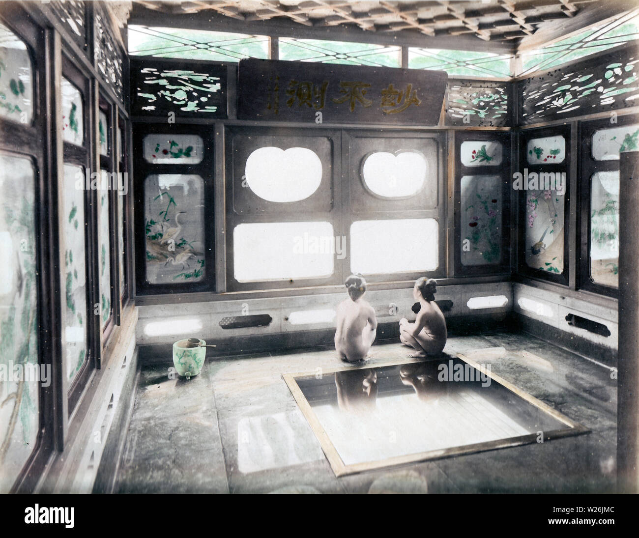 [ 1890s Japan - Japanese Women in Bath ] —   Two women are squatting next to a bath in a beautiful wooden bathroom in Hakone, a famous onsen resort. The windows feature artwork and the panels above them contain gorgeous displays of woodwork. A wooden ladle rests on the marble water pail in the left corner. The photographer's reflection, and that of his camera, is visible above the two birds on the left window.  19th century vintage albumen photograph. Stock Photo