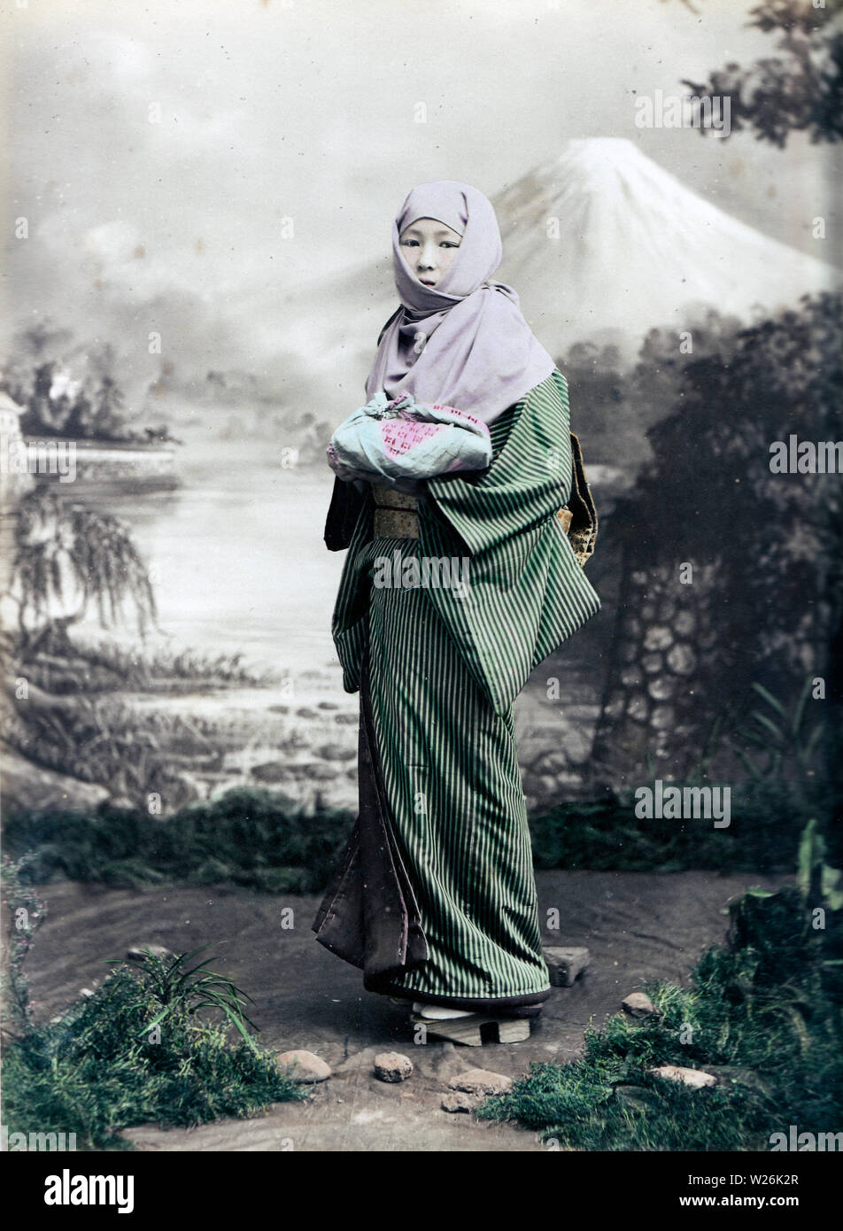 [ 1890s Japan - Japanese Woman in Winter Clothes ] —   A woman in a thick winter kimono has an okosozukin wrapped around her head to protect herself against the winter cold. In her hands she holds something wrapped in a furoshiki, a Japanese wrapping cloth. She is wearing geta and her hair is done in a traditional manner.  19th century vintage albumen photograph. Stock Photo