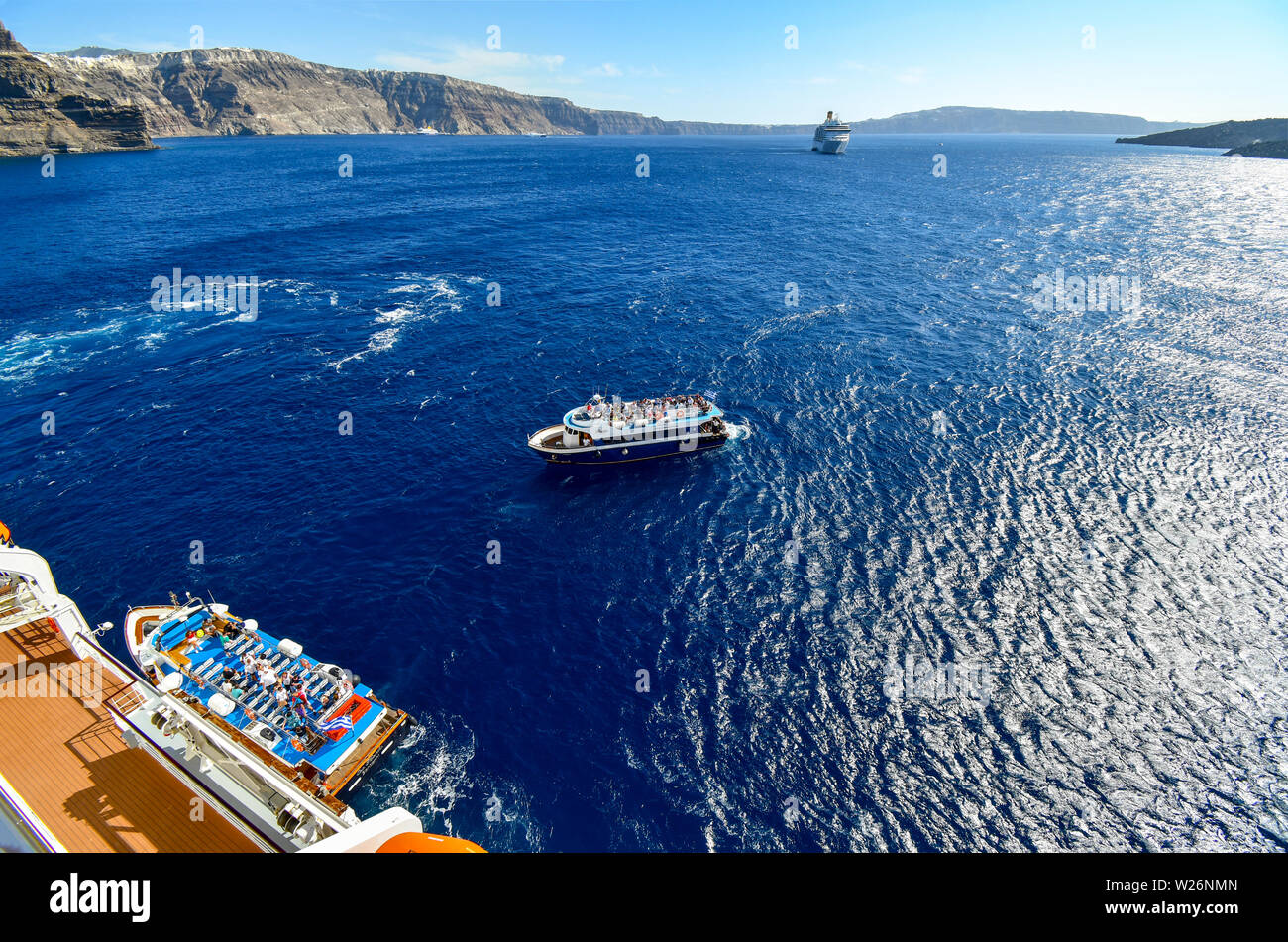 A tender boat pulls alongside a massive cruise ship with another boat and ship in the distance inside the caldera of Santorini Greece Stock Photo