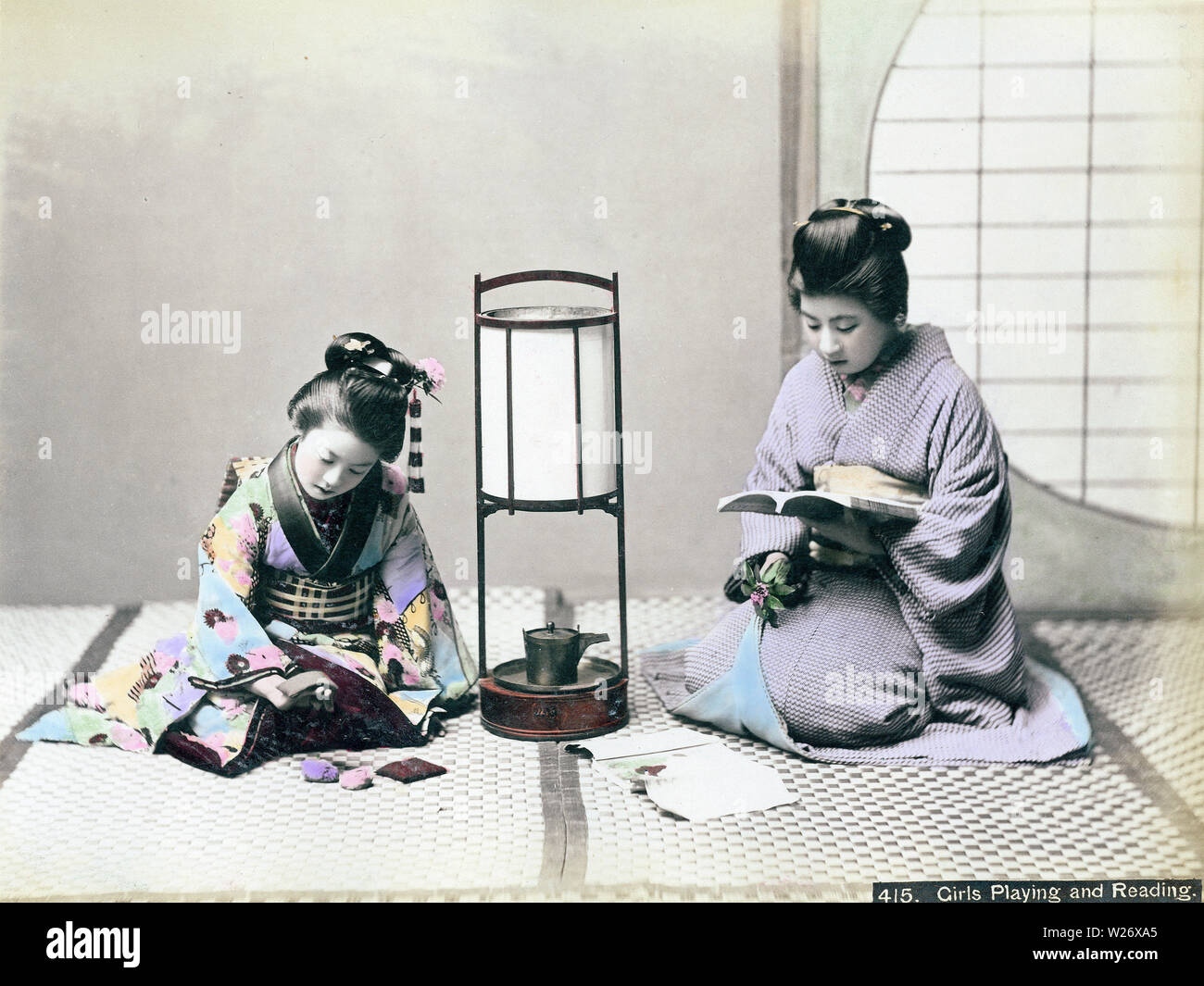 [ 1890s Japan - Japanese Women in Kimono ] —   Two women in kimono reading and playing. An andon lamps stands between them.  19th century vintage albumen photograph. Stock Photo