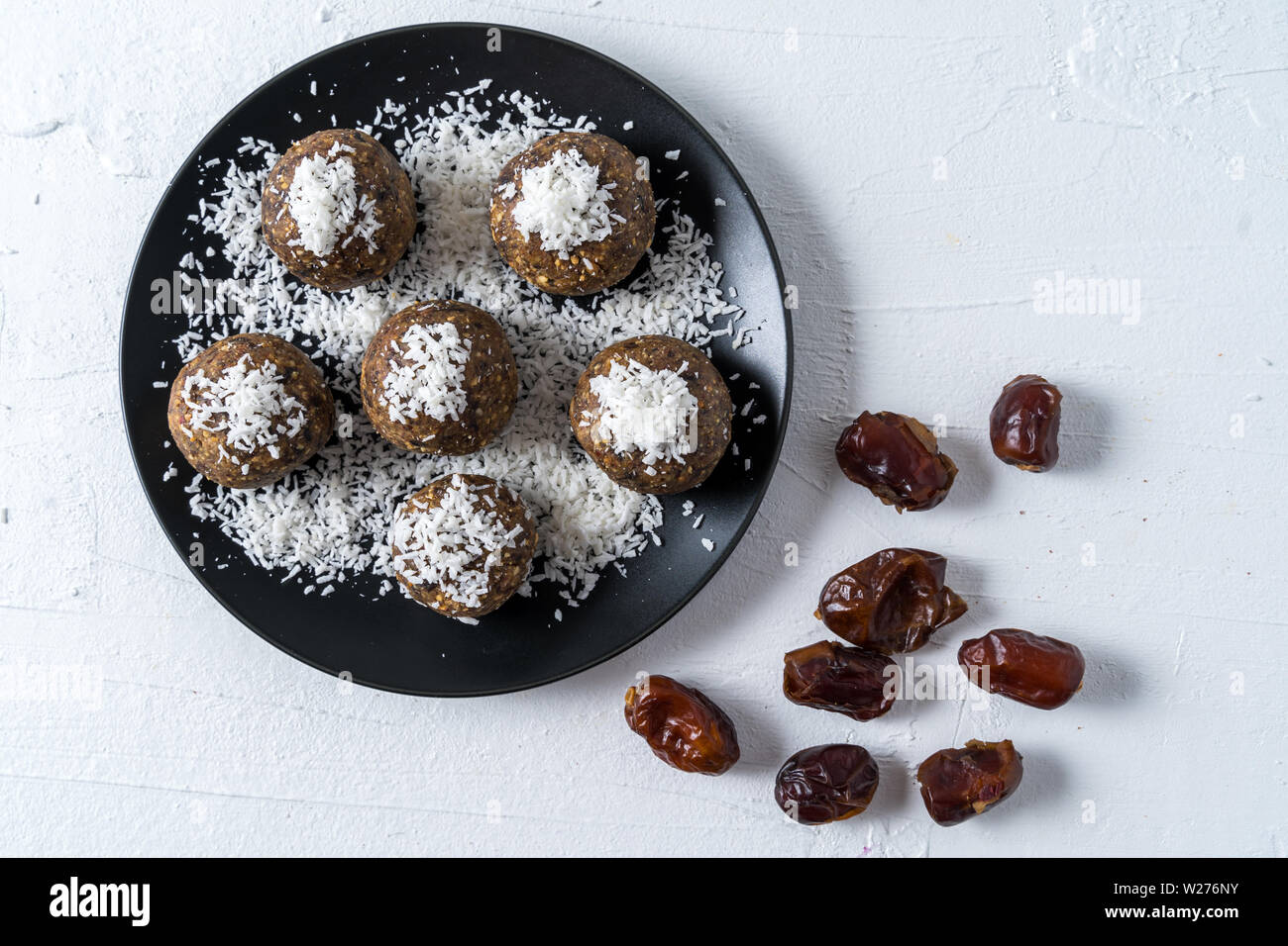 Healthy organic energy balls made with dates, prunes, raisins, peanut, with coconut shavings, in black plate on white background, flat lay. Stock Photo