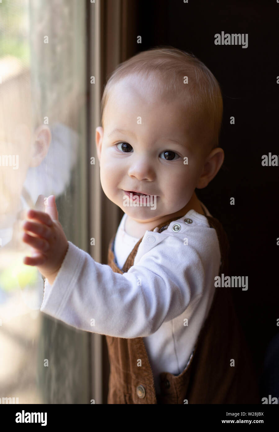Toddler/Baby in overalls standing next to window, looking at camera Stock Photo