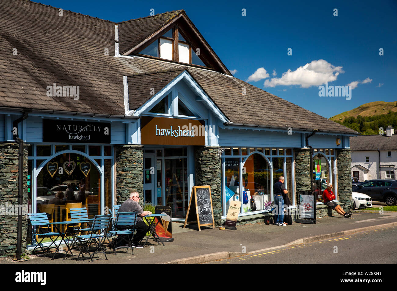 UK, Cumbria, Hawkshead, Hawkshead outdoor clothing shop and Naturally Hawkshead cafe with customer sat in sunshine at outside table Stock Photo