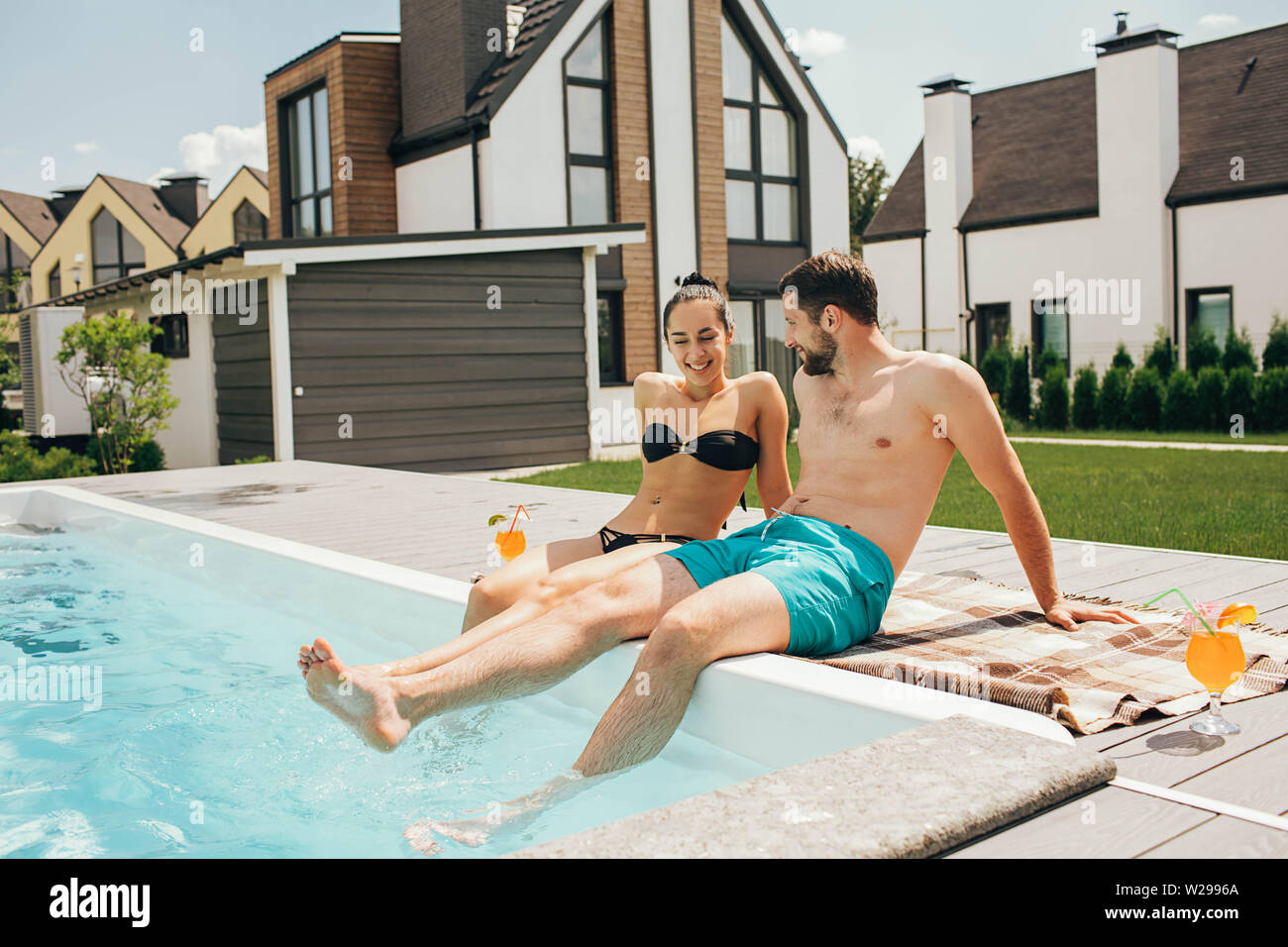 Happy couple sitting by the poolside in the backyard of their house. Man and woman sitting near water pool and funny splashing water their legs Stock Photo