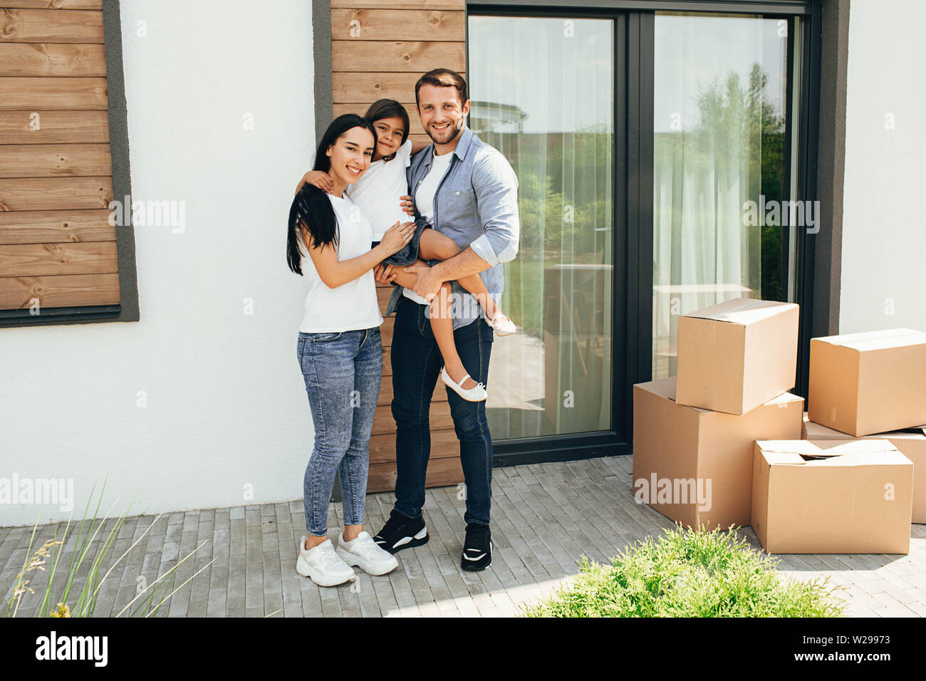 Happy parents with daughter standing on backyard with cardboard boxes. buying an apartment. Stock Photo