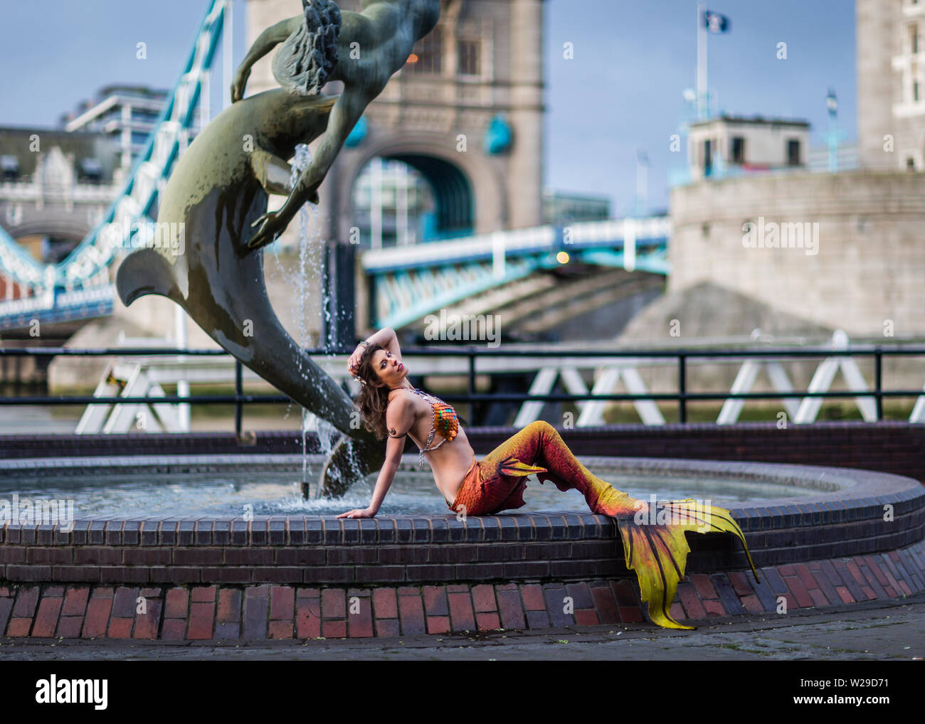 A mermaid poses by the Girl and Dolphin by Tower Bridge in London England. 2 Stock Photo