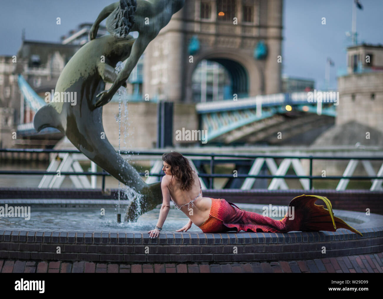 A mermaid poses by the Girl and Dolphin by Tower Bridge in London England. 1 Stock Photo
