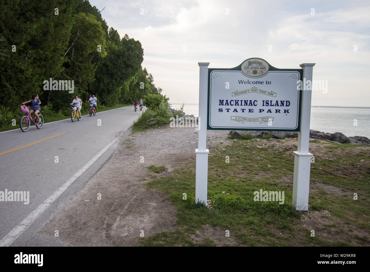 Mackinaw Island, Michigan, USA - Cyclists on a two lane highway around perimeter of Mackinac Island with state park sign in the foreground. Stock Photo