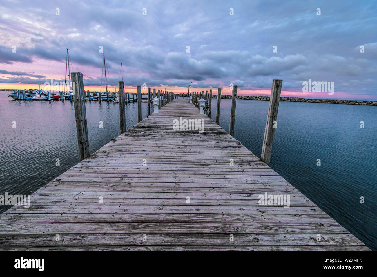 Wooden Dock Sunset. Wooden dock disappears to the horizon with a sunset sky at a harbor marina with various boats in the background. Stock Photo