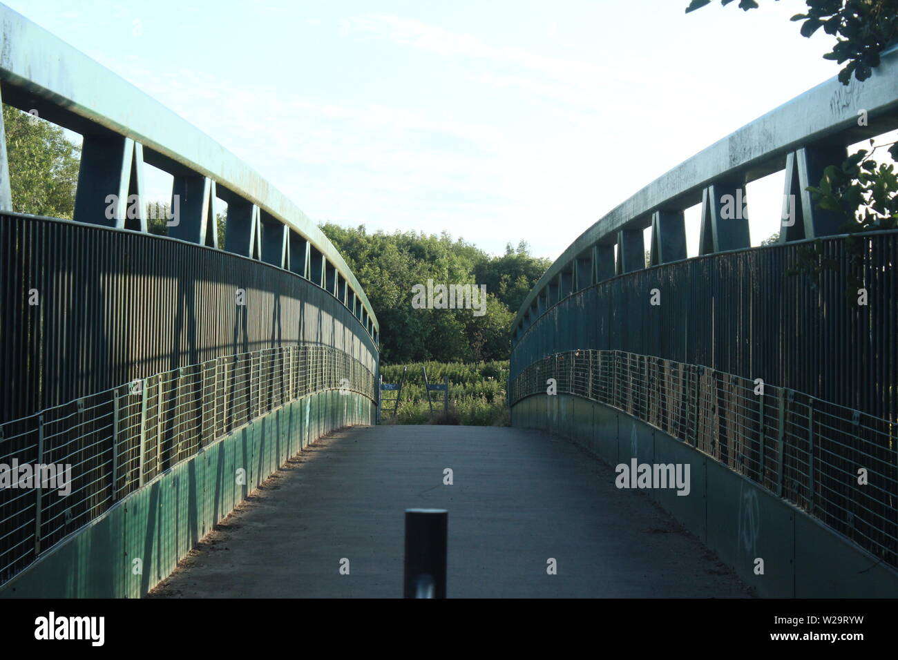 Image of green walkway bridge with bushes and trees in the distance over a lake on a sunny, clear day Stock Photo