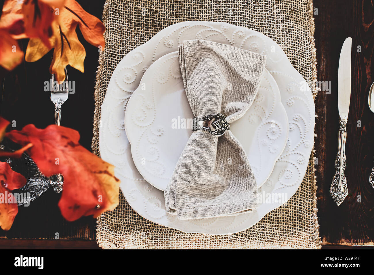 Thanksgiving Day or autumn place setting with napkins and cutlery on burlap table runner over a rustic farmhouse table background. Colorful fall leave Stock Photo