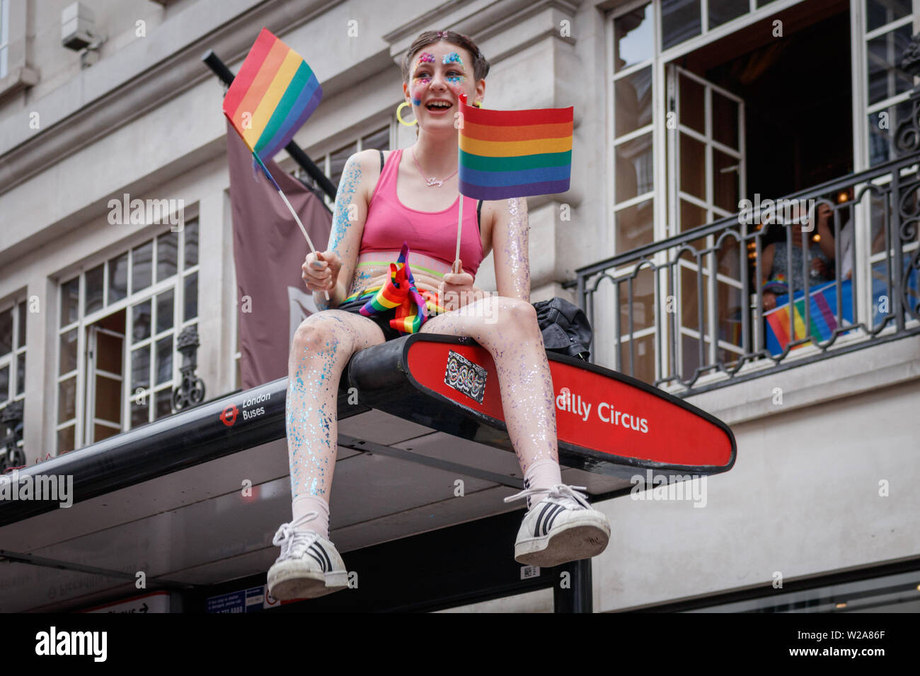 A girl watches from a bus stop, while waving rainbow flags during the parade.The Pride in London 2019 Parade Day is a London-based event held by Pride in London. It is a parade of people of all genders, sexualities, races, nationalities, backgrounds and faiths coming together for the UK’s biggest Pride celebration. Central London was covered in rainbow-themed decorations, signifying inclusiveness. Stock Photo
