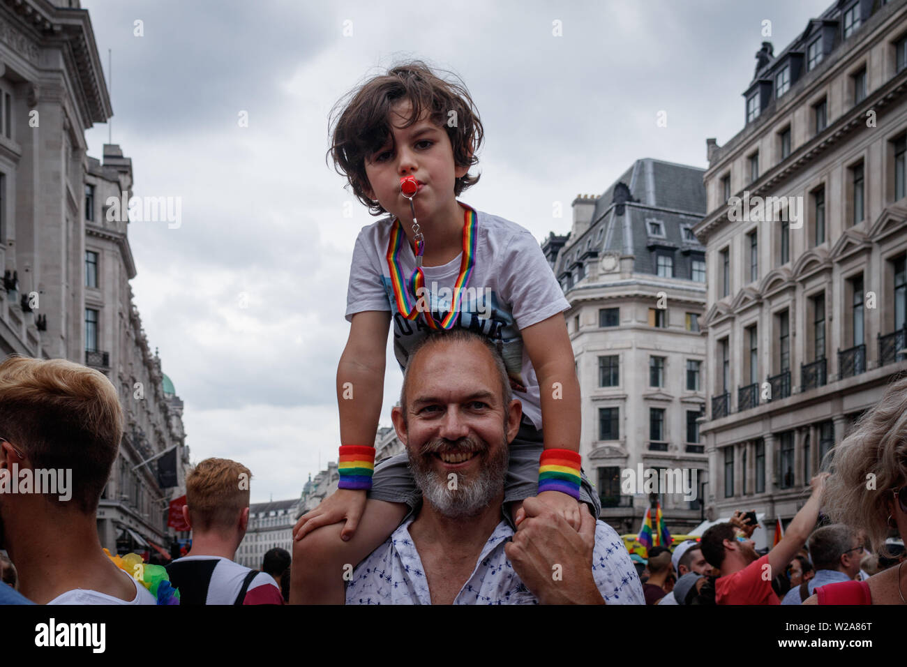 A kid on top of the father's shoulders, with rainbow colours, blows a whistle, during the parade.The Pride in London 2019 Parade Day is a London-based event held by Pride in London. It is a parade of people of all genders, sexualities, races, nationalities, backgrounds and faiths coming together for the UK’s biggest Pride celebration. Central London was covered in rainbow-themed decorations, signifying inclusiveness. Stock Photo