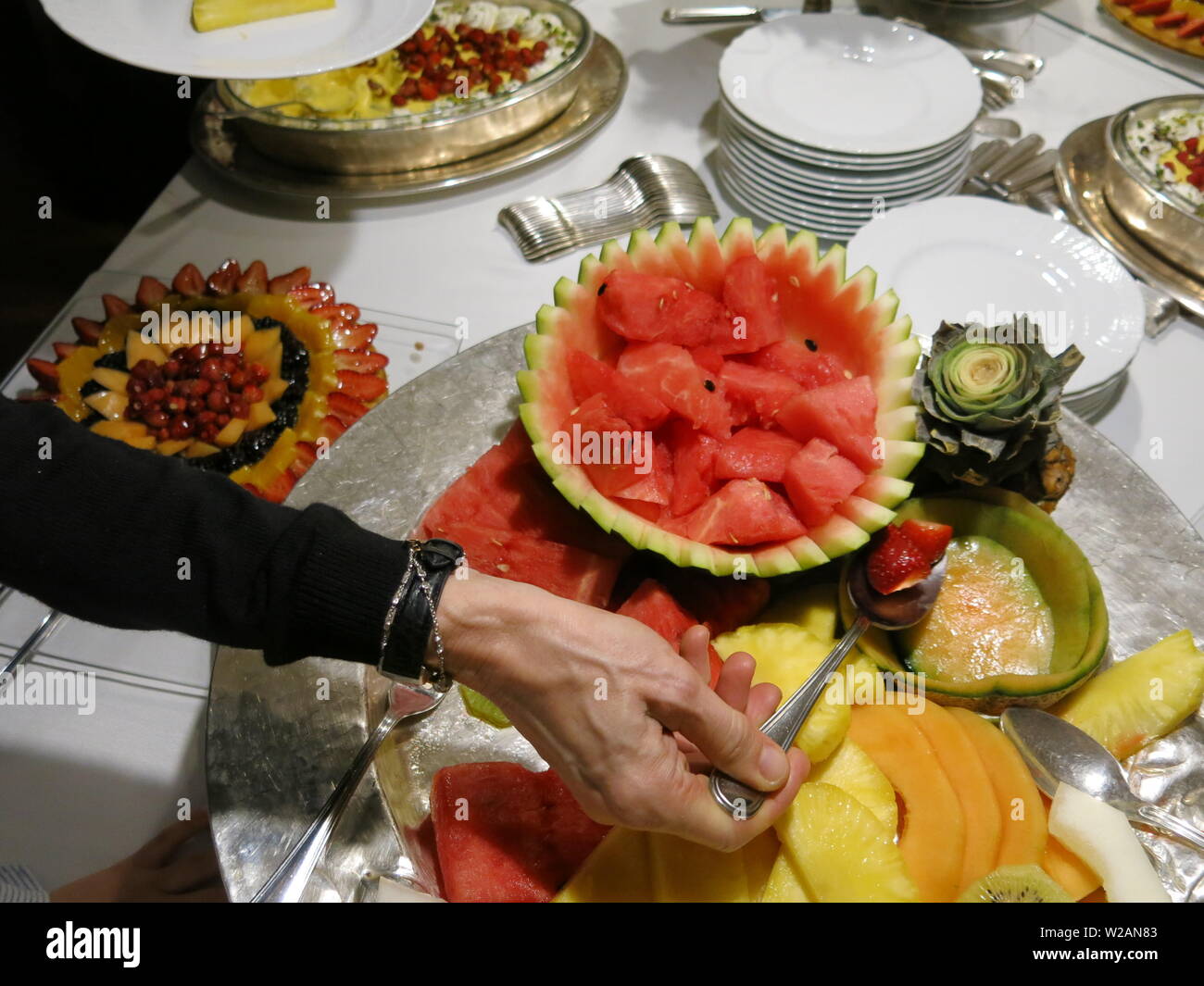A woman's hand is holding a spoon to serve strawberries from an array of desserts and platters of fresh fruit on offer at an appetising buffet Stock Photo