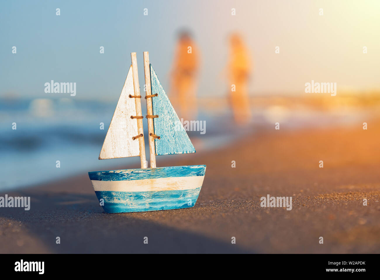 wooden toy sailboat standing on sand near the seashore in summer. Blurred people and toy sailboat at the beach. Vacation and leisure concept concept. Stock Photo