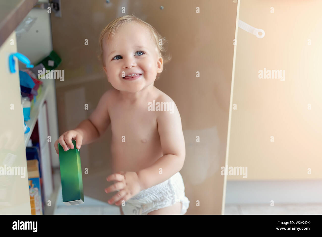 Cute curious caucasian baby boy open cupboard door in kitchen and