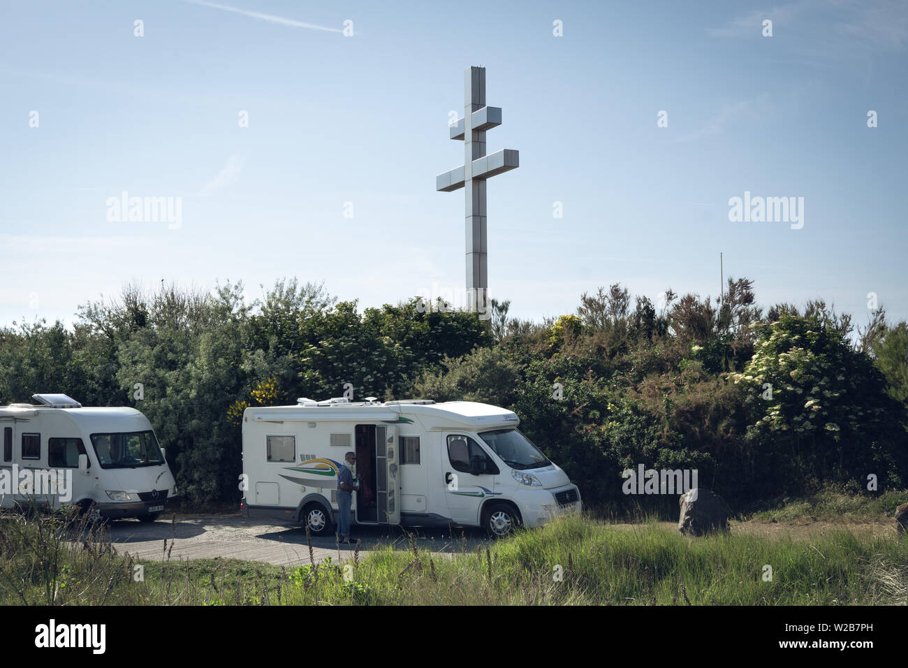 Two RVs (recreational vehicles) parked right behind the Cross of Lorraine, in Graye-Sur-Mer  a man climbing into one the RV can be seen Stock Photo