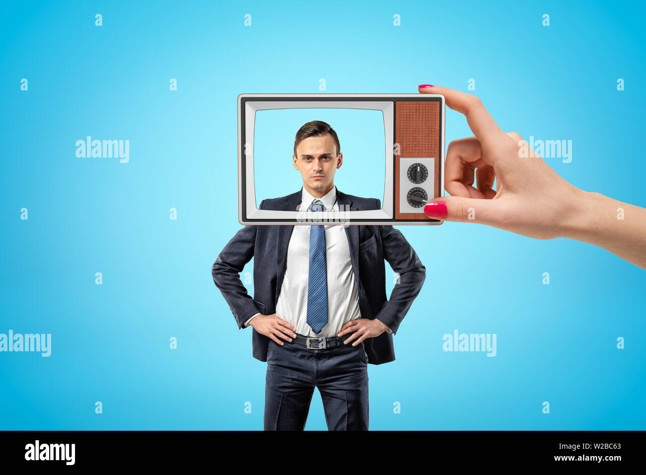 Young serious businessman standing, hands on hips, looking at camera through old TV frame with removed screen, held in woman's hand. Stock Photo
