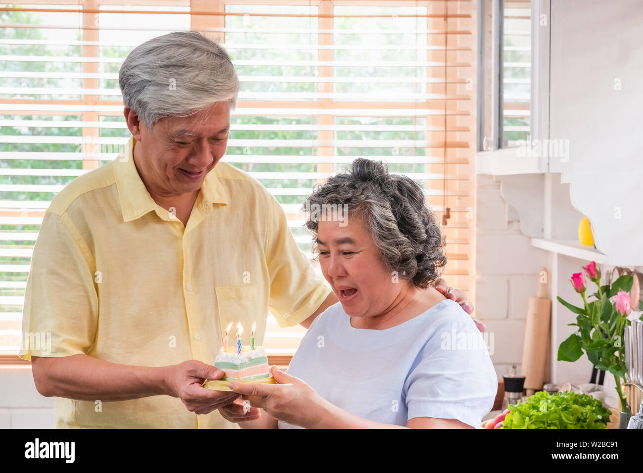 asian senior man surprise senior woman with birthday cake in kitchen at home.aging at home concept Stock Photo
