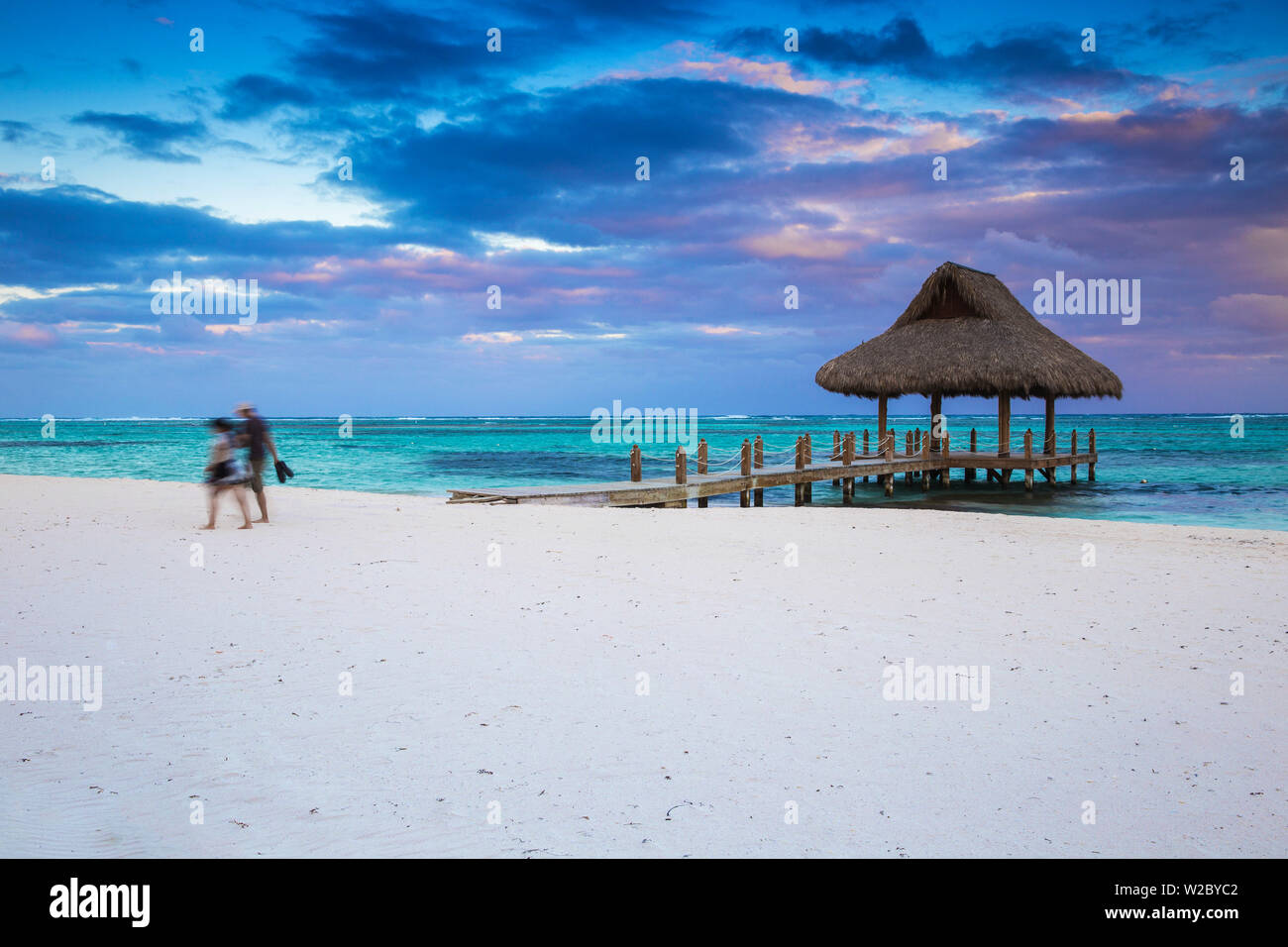 Dominican Republic, Punta Cana, Playa Blanca, Couple walking along beach past wooden pier with thatched hut Stock Photo