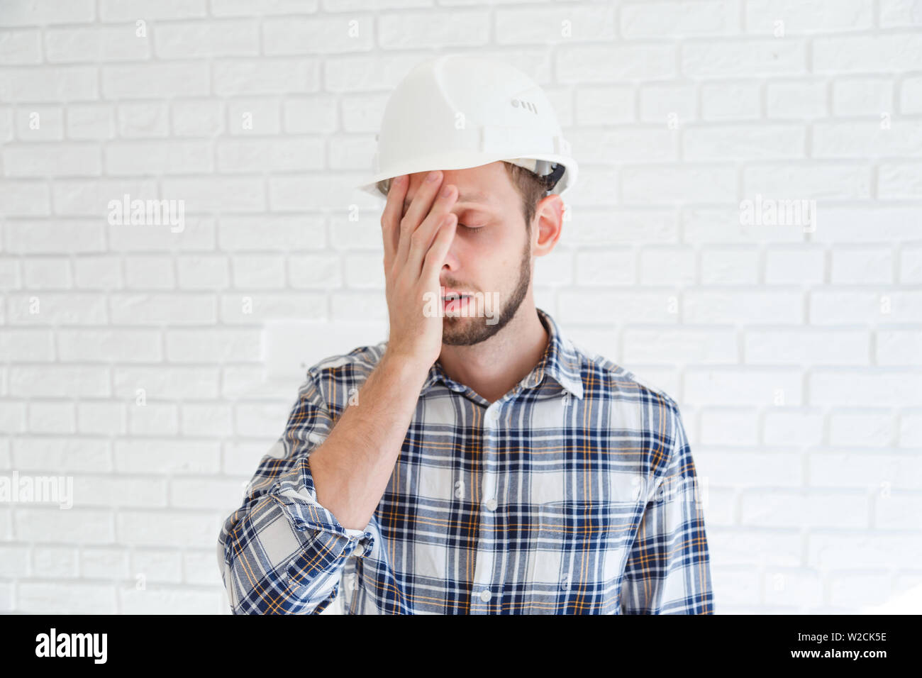 A blue collar worker wearing a hardhat is covering his face with his hand. Stock Photo