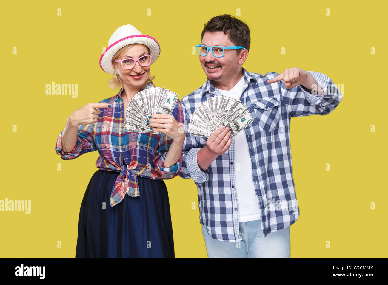 Couple of wealthy friends, adult man and woman in casual checkered shirt standing together holding fan of dollars and pointing fingers, toothy smile, Stock Photo