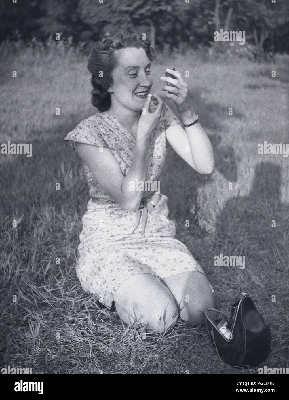 1950s, historical, a young lady in a dress kneeling down outside on a some grass using a small pocket mirror to apply her lipstick, England, UK. Stock Photo