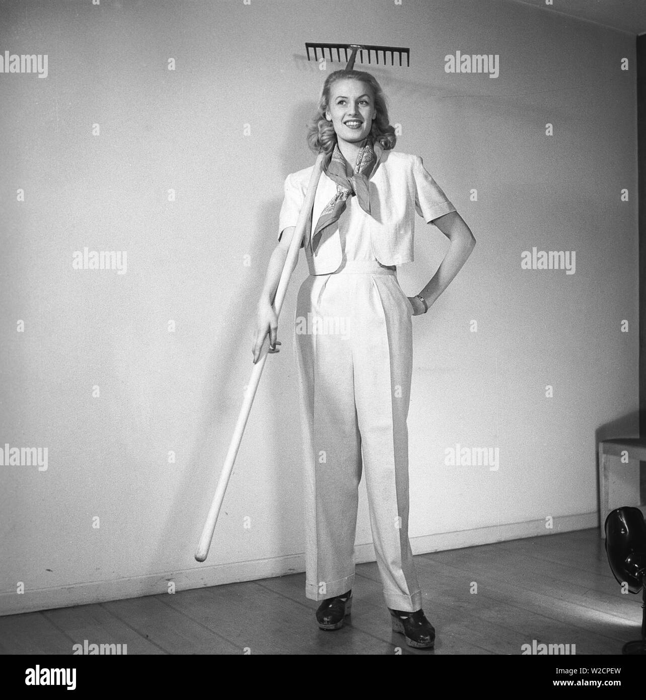1940s summer outfit. A young blonde woman wears a practical summer outfit with long white trousers and a short jacket. By holding a rake it's implied that she is dressed for garden work.  Sweden 1947 Kristoffersson Z25-3 Stock Photo