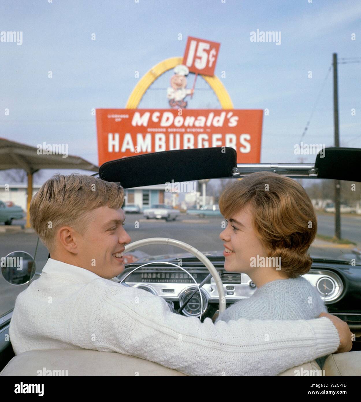 Fast food in the 1950s. A young couple in their convertible car with a McDonald's sign in the background. The yellow arch being part of the original design called the Golden Arches was first used 1953.  The cost of a limited menu hamburger, shake and fries was 15 cents. 1950s 1960s ref 5-40-4 Stock Photo
