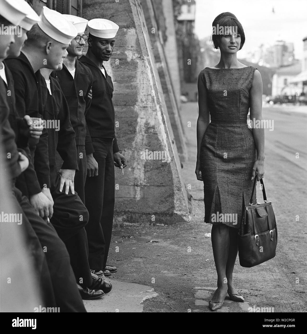 Fashion in the 1960s. A group of American sailors are standing at the docks on shore leave and makes a nice addition to the fashion shoot. A young model passes them wearing a dress with matching handbag and shoes. Sweden 1964 ref BV59-4 Stock Photo