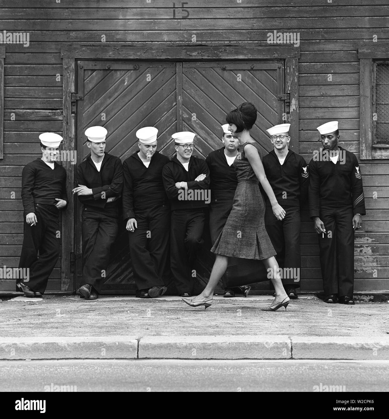 Fashion in the 1960s. A group of American sailors are standing at the docks on shore leave and makes a nice addition to the fashion shoot. A young model passes them wearing a dress with matching handbag and shoes. Sweden 1964 ref BV59-5 Stock Photo