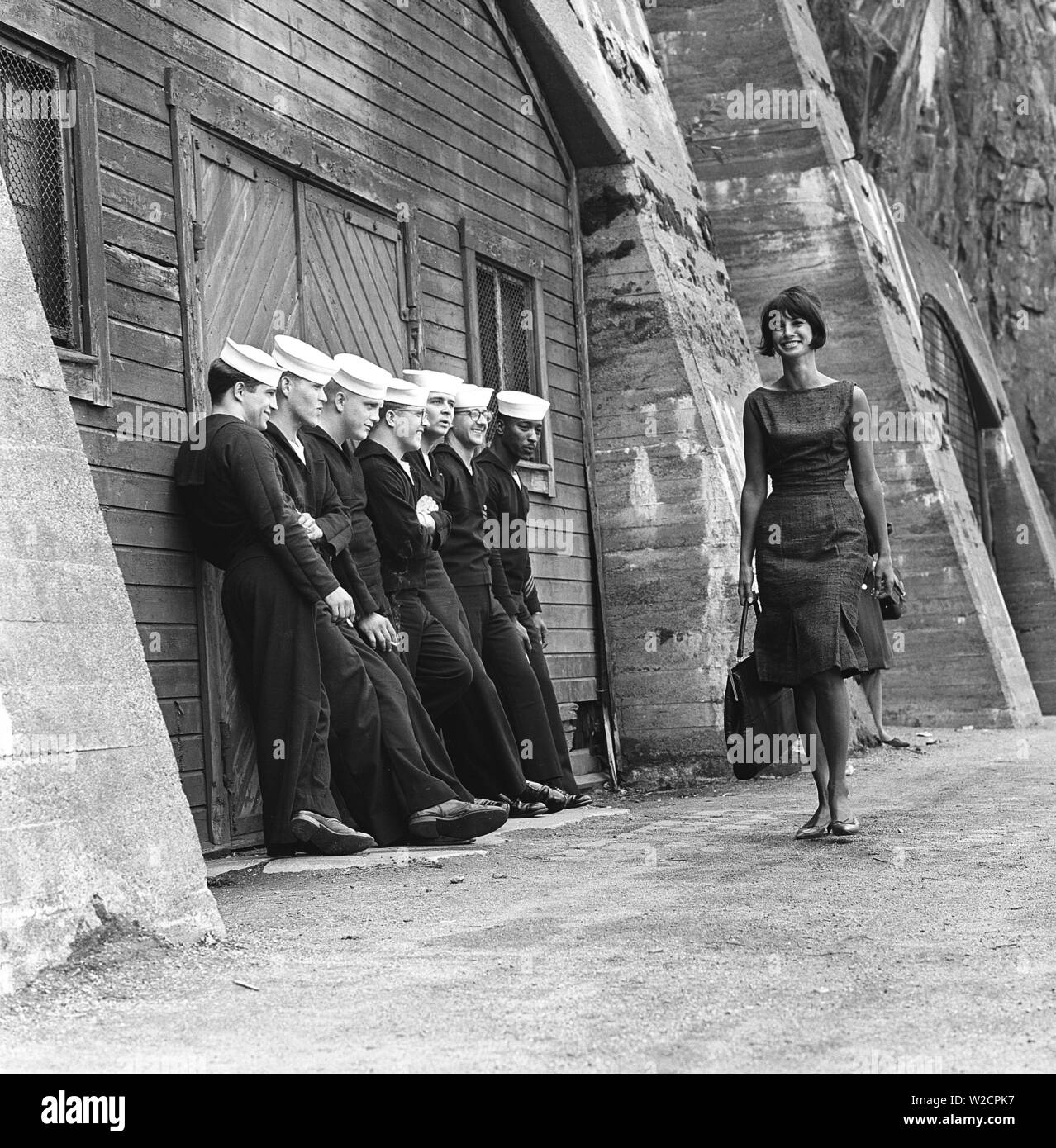 Fashion in the 1960s. A group of American sailors are standing at the docks on shore leave and makes a nice addition to the fashion shoot. A young model passes them wearing a dress with matching handbag and shoes. Sweden 1964 ref BV59-6 Stock Photo