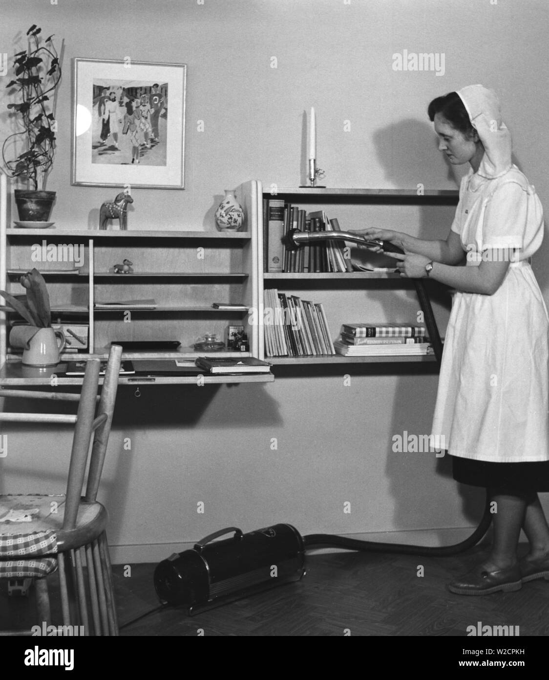 Vacuuming in the 1950s. The young student Gertrud Larsson is having a cleaning day in her student apartment. She is thorough and even the books in the bookshelf gets a good vacuum cleaning. Sweden 1954 Stock Photo