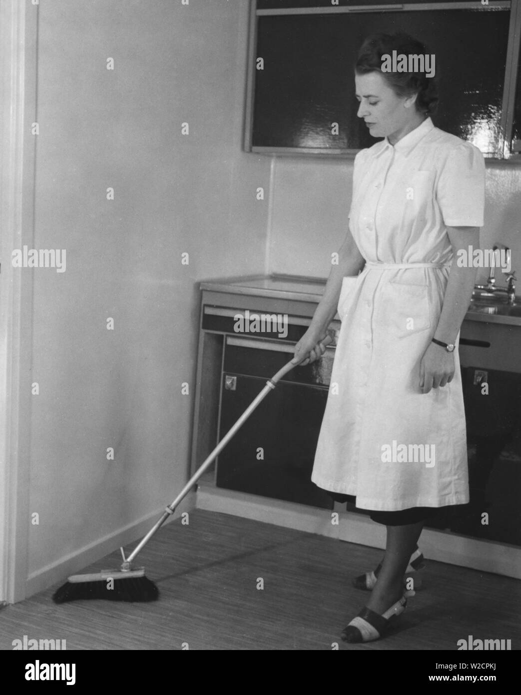 Cleaning day in the 1950s. A young woman dressed in a practical cleaning outfit,  is cleaning the kitchen and is using a brush with an telescoping handle. A new invention to stand up straight when dusting and avoid back pains. Sweden 1950 Stock Photo