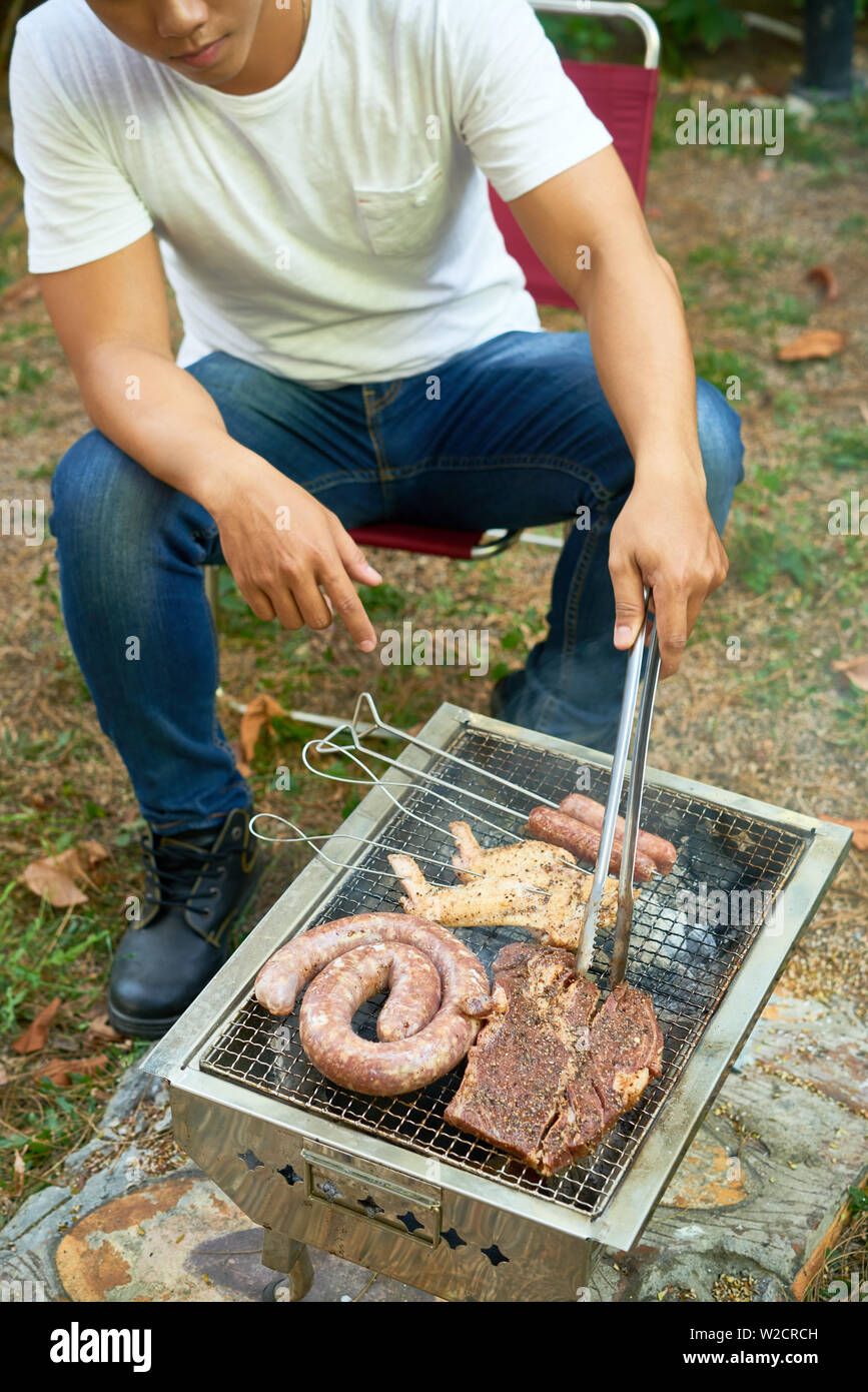 Young man sitting outdoors and cooking meat and sausages on the grill Stock Photo