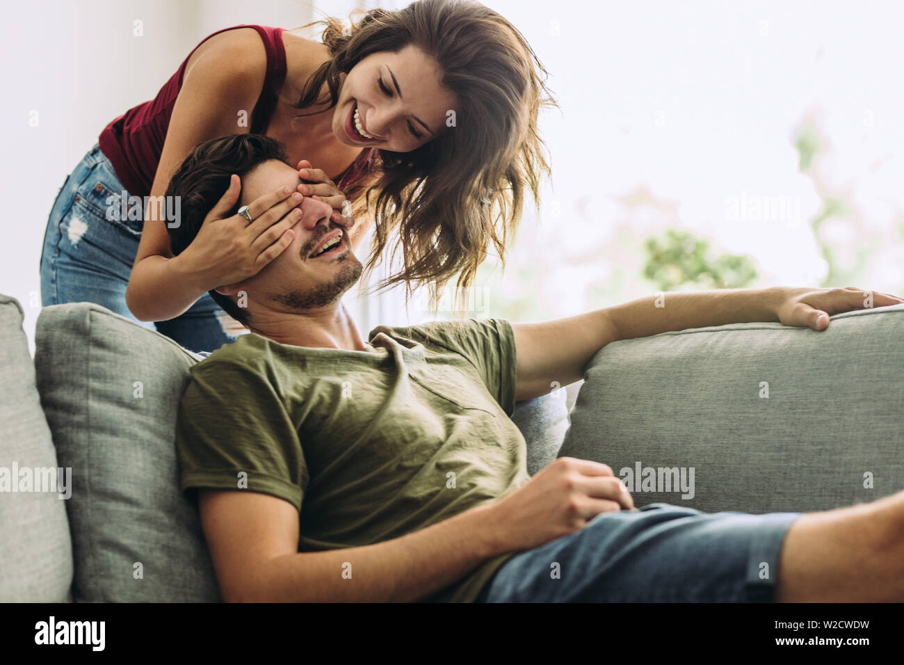 Young woman standing behind her boyfriend on sofa and covering his eyes at home. Woman surprising man at home. Stock Photo