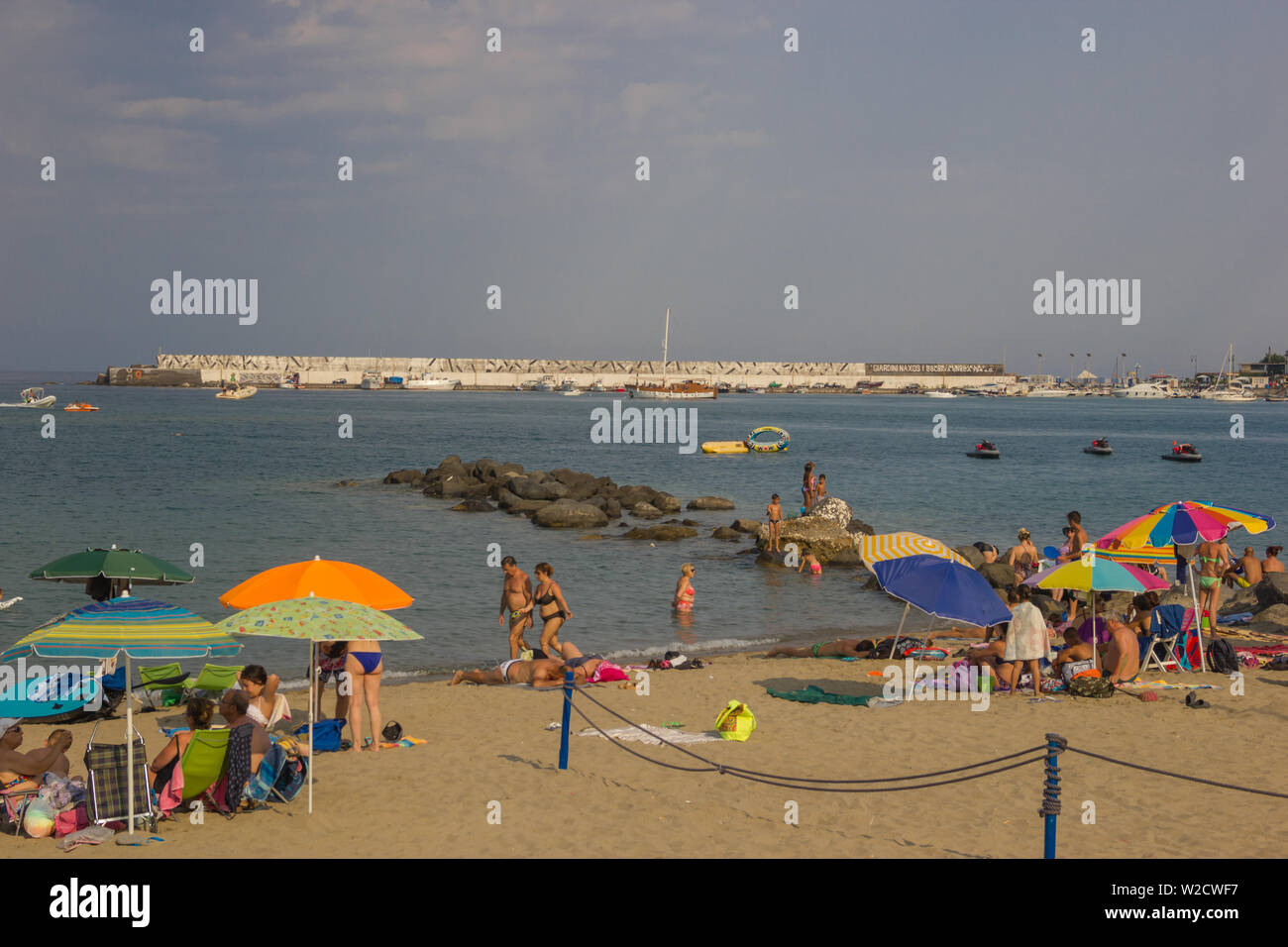 Giardini Naxos Sicily Italy 2019 Summer view of the beautiful beach with people enjoying the sun and the sea, with the far tourist harbor Stock Photo