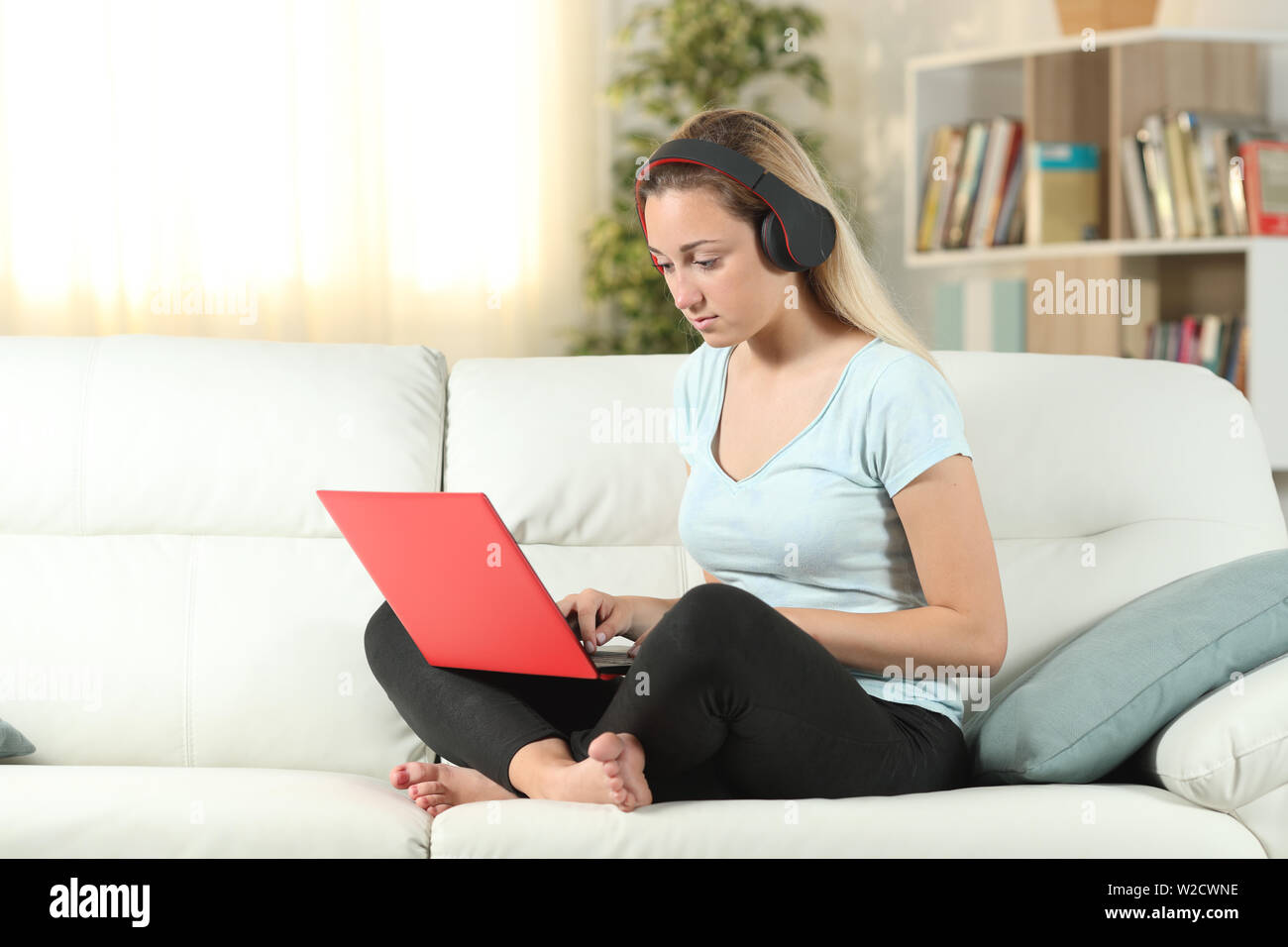 Serious girl using a laptop and headphones sitting on a couch in the ...