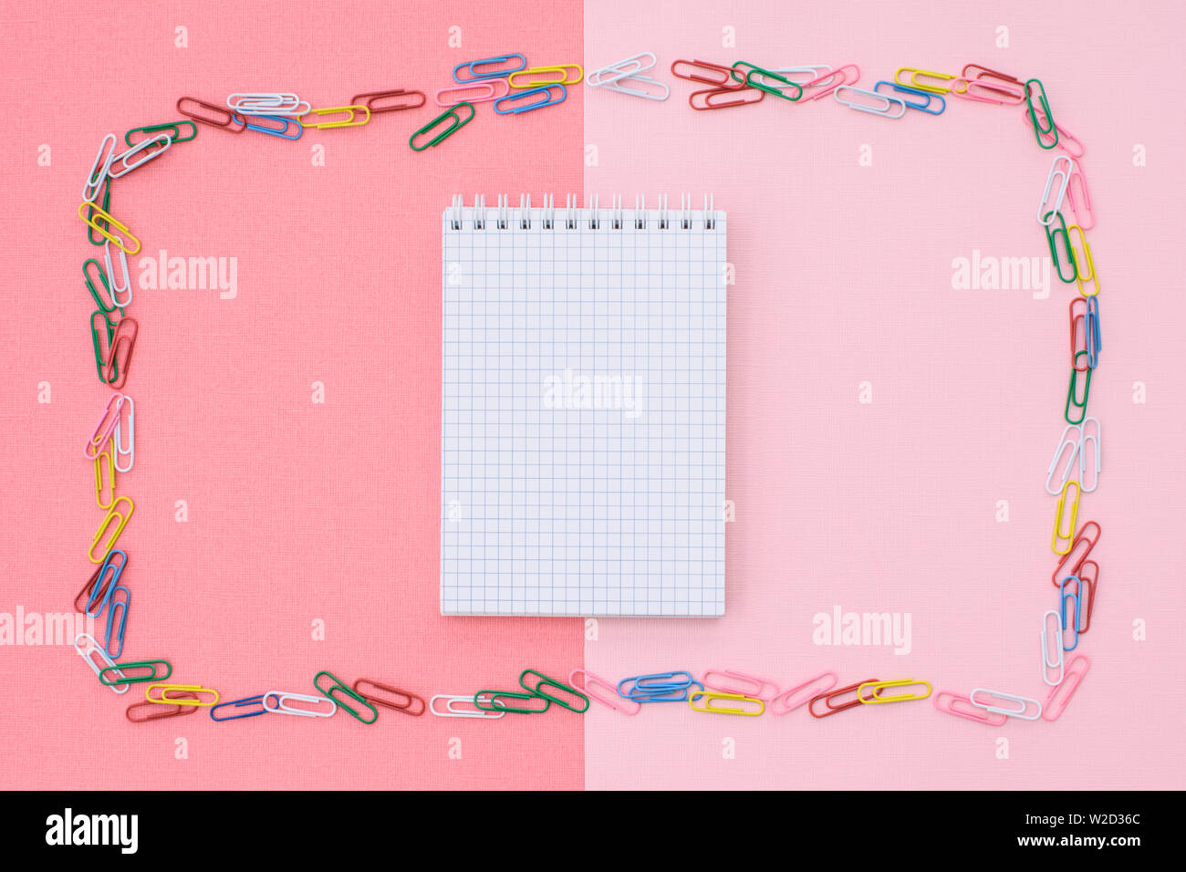 Notebook in cage in a frame of colored paper clips on a double background - pink and coral. Concept back to school. Stock Photo