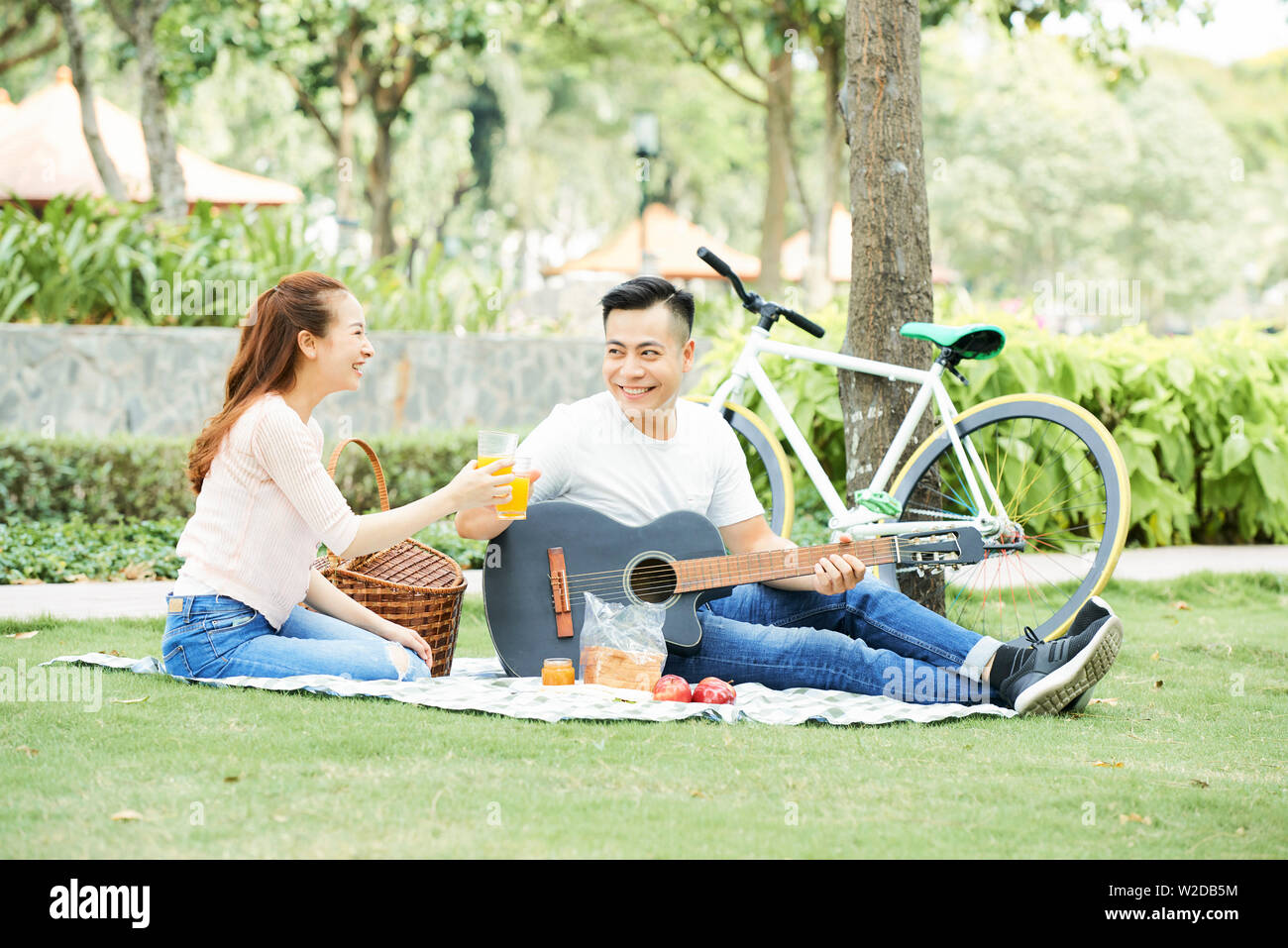 Happy young Asian couple sitting together in the park playing guitar ...