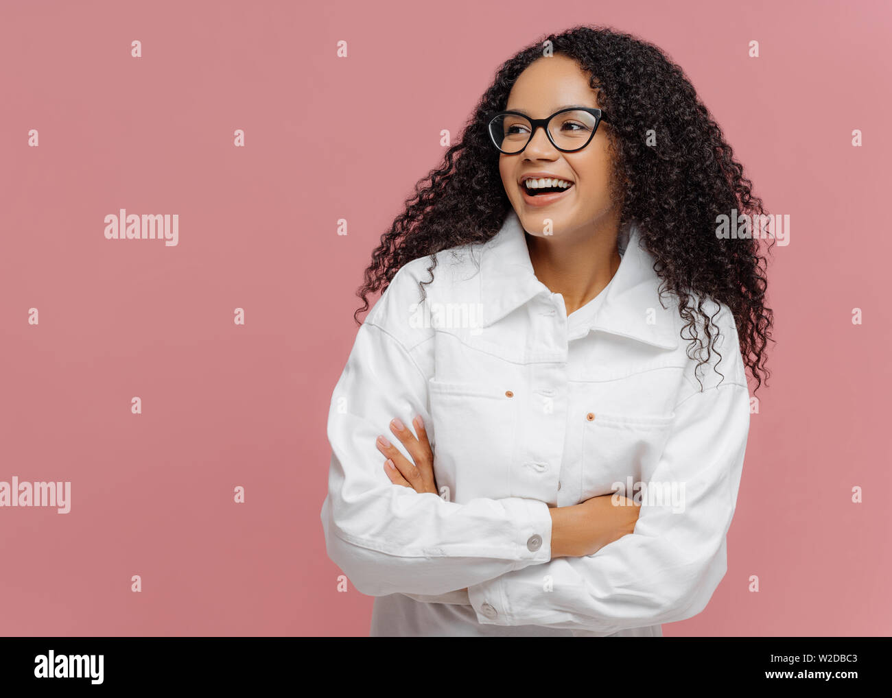 Carefree cheerful Afro American woman wears white jacket, keeps arm folded, looks aside, has charming smile, enjoys nice day in company of friends, st Stock Photo