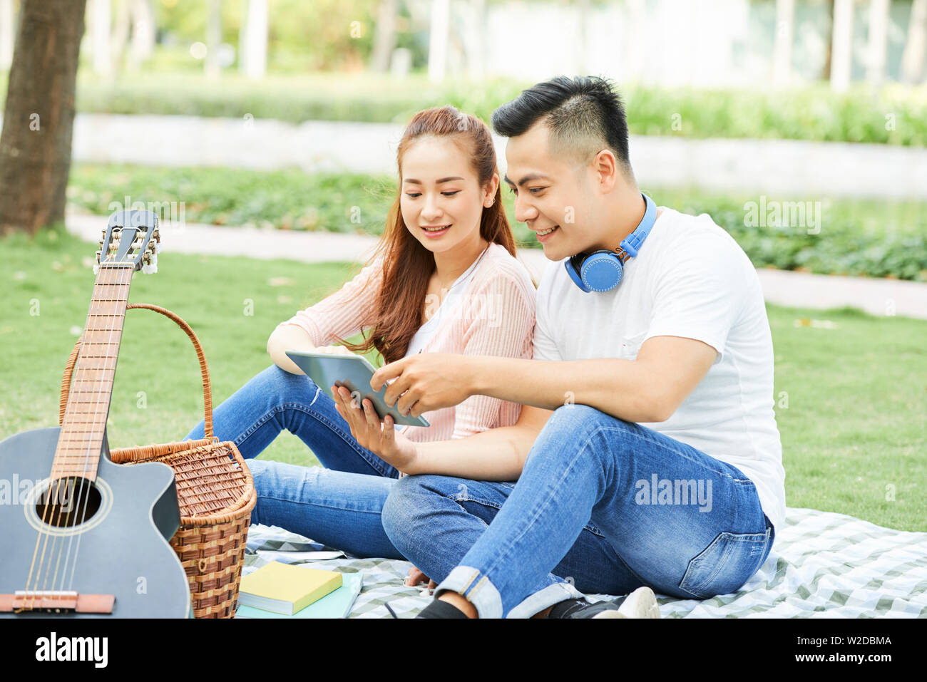 Asian happy couple sitting on the grass and communicating online using digital tablet during their date in the park Stock Photo