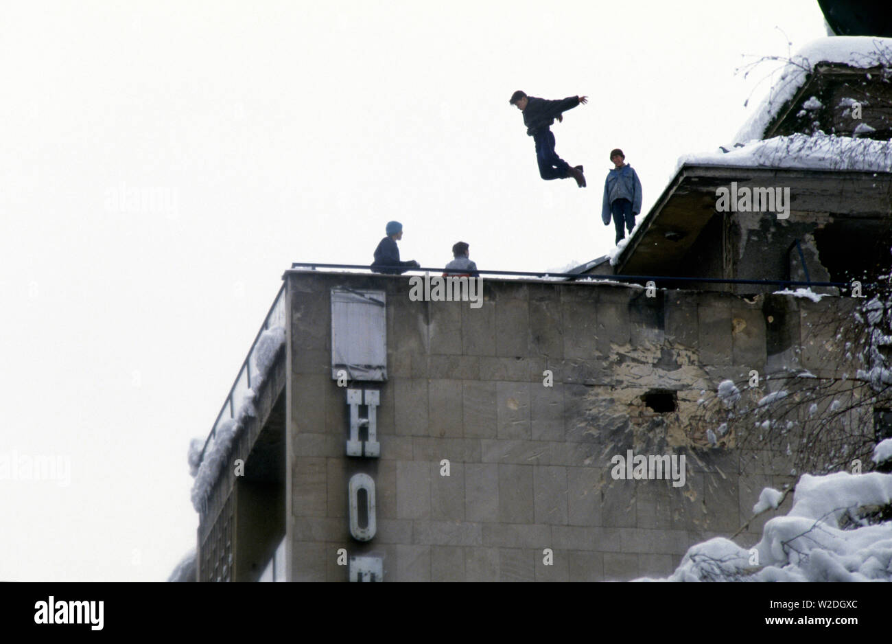 28th March 1993 During the Siege of Sarajevo: young boys play on the roof of the Hotel Europa, high above Jugoslovenske Narodne Armije (renamed Zelenih beretki Street after the war). Below them is a big hole in the wall, caused by a mortar bomb or artillery shell. Stock Photo