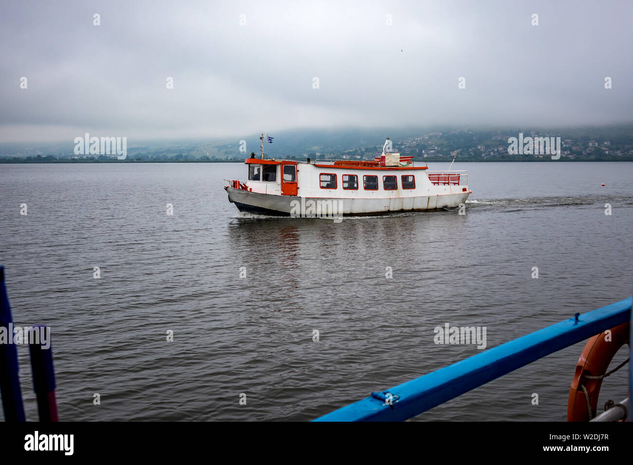 Lake Ioannina, Greece, small rusty old white and red ferryboat sailing in the waters of the lake between the island and the beautiful Greek town. Foggy spring morning Stock Photo